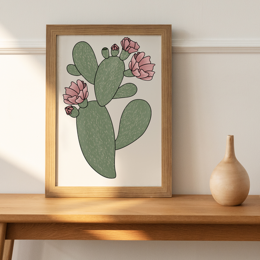 Prickly pear cactus with several pink flowers in various stages of bloom in an oak frame upon a desk